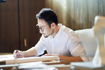 Asian bearded designer sitting, thinking and sketching ideas indoors