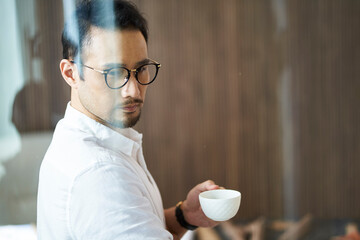 confident Asian businessman standing behind glass drinking coffee looking down at the city