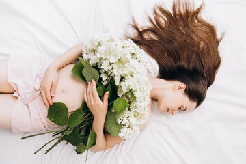 Obraz na płótnie Canvas Future mother's expectation. Head shot portrait young pregnant woman with perfect skin touching gently tummy hand lying on comfortable bed with beautiful fresh summer flowers. Concept motherhood