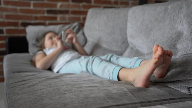 A little girl lies at home on the couch and looks into a smartphone. The child plays and watches the video. The girl has bare feet. The child is wearing pajamas. 4K. Focus on legs