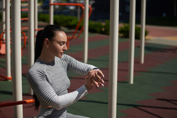 Young sportswoman checking her fitness tracker, pulse and heart rate, lost calories during outdoor workout, standing on a sports ground