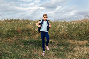 Little girl with a backpack walks among the grass. The child travels on foot with a backpack