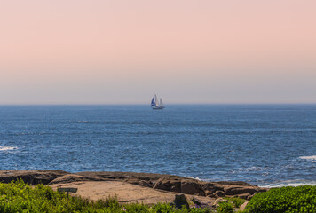 A sailing boat can be seen from a beachside in Maine USA. Beachside is overlooking the ocean and orange sky.