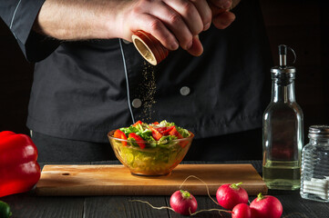 Chef in a restaurant kitchen adds peppers to a fresh vegetable salad. Close-up of a cook hands holding mill. Cooking healthy and tasty food