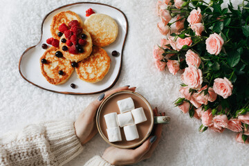 Woman holding a cup with cocoa and marshmallows, flowers and a plate with cheesecakes and berries. Close up. Breakfast concept
