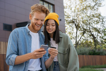 Couple of smiling stylish friends holding credit card, using mobile phone for online shopping. Asian woman and caucasian man ordering food 