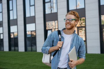 Portrait of happy confident man wearing eyeglasses looking away, copy space. Smiling university student with backpack and laptop computer walking in university campus, education concept 