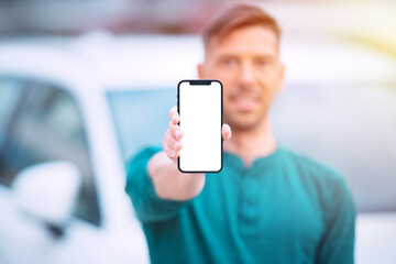 Smartphone mockup, blank screen. Young smiling successful man standing near white car outdoors and showing or holding his modern phone with blank space