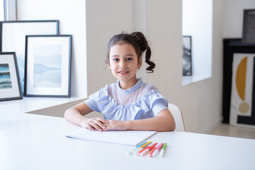 Naklejka premium A beautiful young schoolgirl is drawing at a white table in her room, looking at the camera smiling. home learning concept
