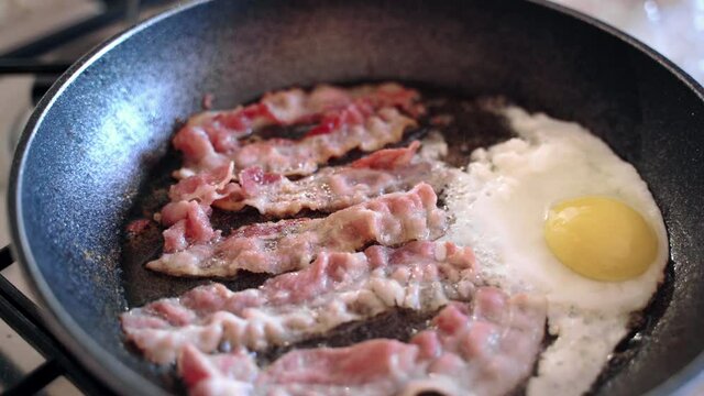 Top view of round pan on gas stove with sizzling olive oil and frying bacon slices with chicken egg, preparation of morning nutritious breakfast at home. English breakfast consisting of fried bacon