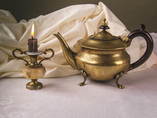 Art Deco silver teapot and romantic candlestick with lit candle on table covered with white tablecloth with folds, evening atmosphere.