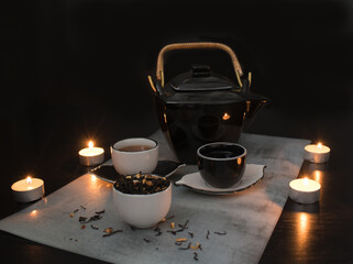 Fototapeta na wymiar Beautiful black and white tea ceremony set: teapot and small cups and saucers next to a cup filled with dried tea leaves with pieces of citrus on the background of a black table with burning candles