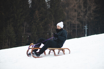 Tobogganing in winter at dizzying speed on the piste with a smile on your face. Kids' joy. A boy in a black jacket and white hat rides on a wooden sledge from times past. Enjoying winter