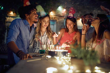 Group of cheerful friends lighting candles on the birthday cake at the open air party. Quality friendship time together