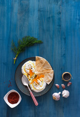 Dish with Turkish eggs Cilbir, plain yogurt, poached eggs, garlic, chilli, fresh dill and melted butter. Embellished with few pieces of lavash bread and black sesame seeds. Dark blue background.