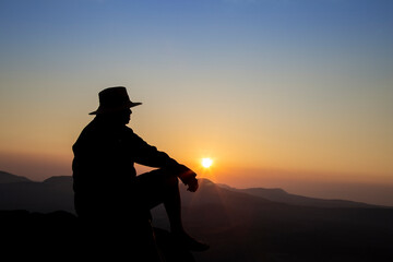 Fototapeta premium Silhouette of old man sitting on mountain peak during sunrise, man enjoying with nature view, landscape