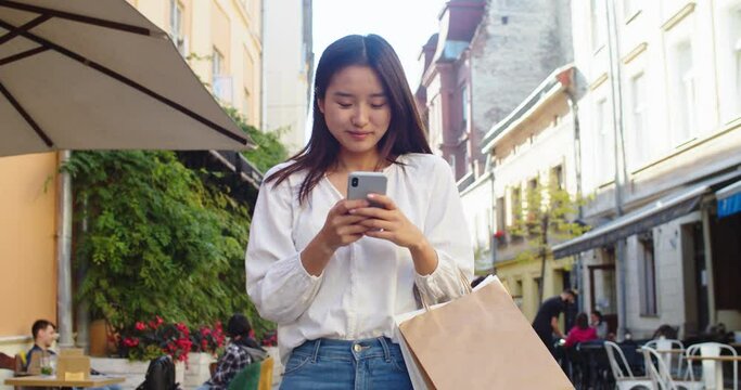 Beautiful lady walking after shopping with purchases, texting and chatting with friends. Young Asian woman with paper bags going in city street and using smartphone to browse, scroll news feed.