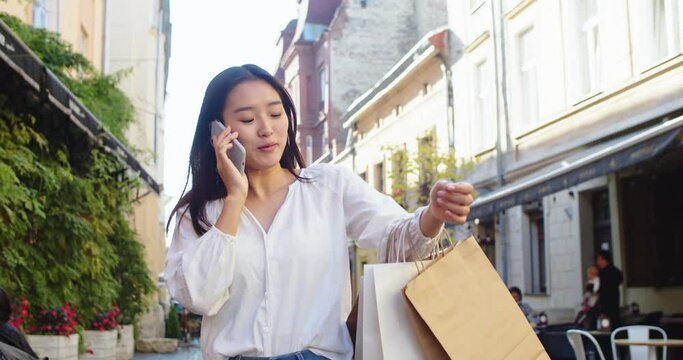 Smiling curly woman wearing trendy sunglasses walks down the central city street and uses her phone. Pretty summer woman in white jacket walks down the street looking at her mobile phone