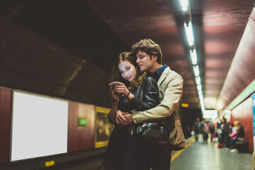 Young beautiful caucasian couple waiting for underground hugging using smartphone surfing web, buying online and watching video