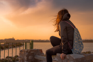 Young brown hair woman wearing a backpack looking at the horizon during sunset near the sea