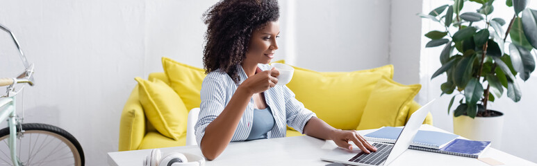 Smiling african american woman with cup of coffee using laptop at home, banner