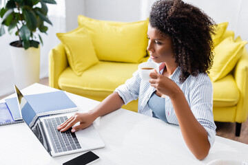 African american teleworker with cup of coffee using laptop with blank screen at home