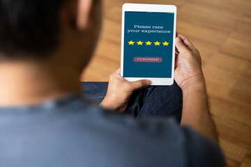 Tablet screen displays a Please rate your experience message to confirm Service satisfaction. A man sits and holding tablet in the work at home. Concept of business service survey. Blurred background