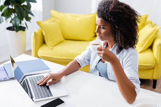 African american teleworker with cup of coffee using laptop with blank screen at home - Powered by Adobe