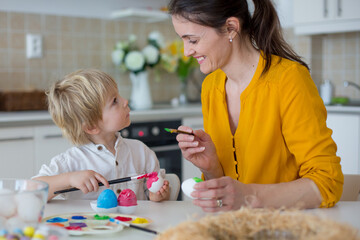 Beautiful blond child, toddler boy, painting easter eggs with mother at home, making easter wreath