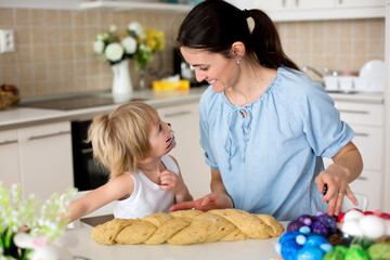 Little blond toddler child with painted face as rabbit and young mother, preparing dough for easter brioce buns, sweet easter bread