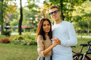 Attractive cute couple is standing in the park, posing for the camera, holding their hands and smiling. A caucasian man and hispanic woman are dating and feeling happy.