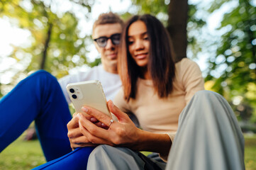 Close up photo of a mobile phone in the woman's hands, focus on device. Surprised couple is using smartphone, watching something on phone, texting, surfing the social media, when sitting in the park.