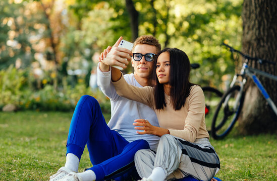 Youthful beautiful couple is taking selfies on a smartphone and sitting on a green grass in the city park. Two attractive people are taking photos on a mobile phone.