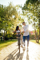 Vertical photo of two people, a couple, in the city park on a sunny day, a woman is sitting on a bicycle, a man is holding her and talking to her. Lifestyle concept.