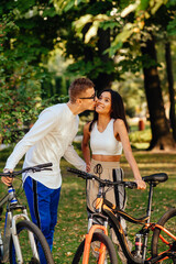 Vertical photo of a lovely couple standing with their bikes in the park. Attractive romantic guy is kissing his girlfriend on the cheek, the woman is smiling and feeling joy.