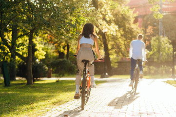 Back view of a female rider on a bright orange bike following her boyfriend bicyclist in a modern city park on a bright summer sunny day.