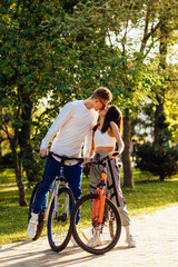 Fototapeta na wymiar Vertical photo of a lovely kissing couple standing outside with their bicycles, feeling romantic in the city park on a sunny summer day. Relationship concept.