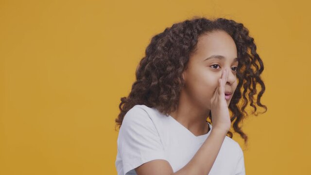 Childhood secrecy. Studio portrait of cute african american curly girl whispering aside, covering mouth with hand