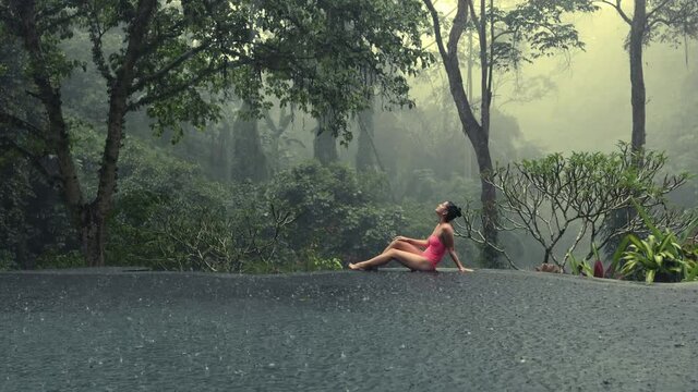 Beautiful young woman in pink bikini enjoying tropical rain falling on her in infinity pool with mystic jungle view