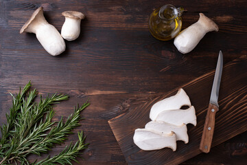 Border from king oyster mushrooms, bottles of olive oil, rosemary sprigs, knife and cutting board on a dark wooden background.