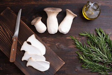 Three raw king oyster mushrooms, rosemary sprigs, mushroom slices on a cutting board, olive oil on...