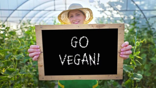 A woman farmer invites buying organic vegetables and fruits. Agriculture. Smiling woman with a sign in her hands calls for healthy food. Vegetarianism, vegan. Greenhouse, family vegetable growing busi