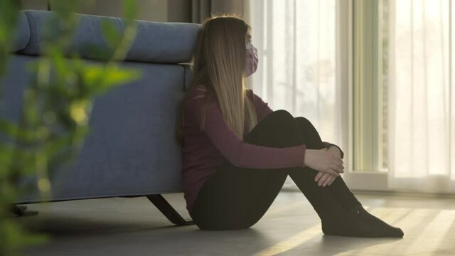 sad girl adolescent in quarantine at home wearing protective mask looking out the window,child feels alone and depressed in isolation during coronavirus covid-19 pandemic,social distancing