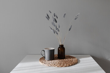 still life with grey cup and dry grey flowers in the brown vase on on the rattan napkin near grey wall. 