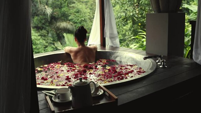 Beautiful young woman relaxing in round outdoor bath with tropical flowers overlooking tropical rainforest jungle in luxury spa