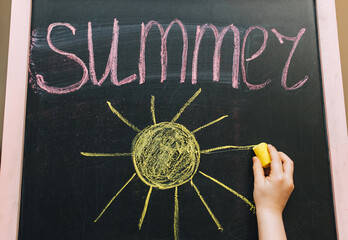 A child, a little girl writes beautifully on a wooden board, an easel, holding yellow chalk in his hand, the word summer and draws the sun. Photo, copy space.