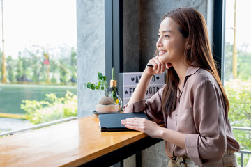 Obraz premium Beautiful Asian woman sit in corner of coffee shop with day light and look outside. She is smiling with happy emotion and think about her small business and sustainable activity.