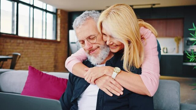 Close uo of affectionate middle-aged couple looking at a laptop together on a sofa in their living room. Husband and wife browsing the internet or social media with a smile
