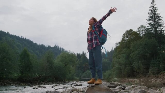Happy girl standing on rock at river bank. Female hiker raising hands in air
