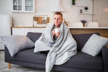 Young man sits on the sofa with a cup of tea wrapped in a blanket and is sick